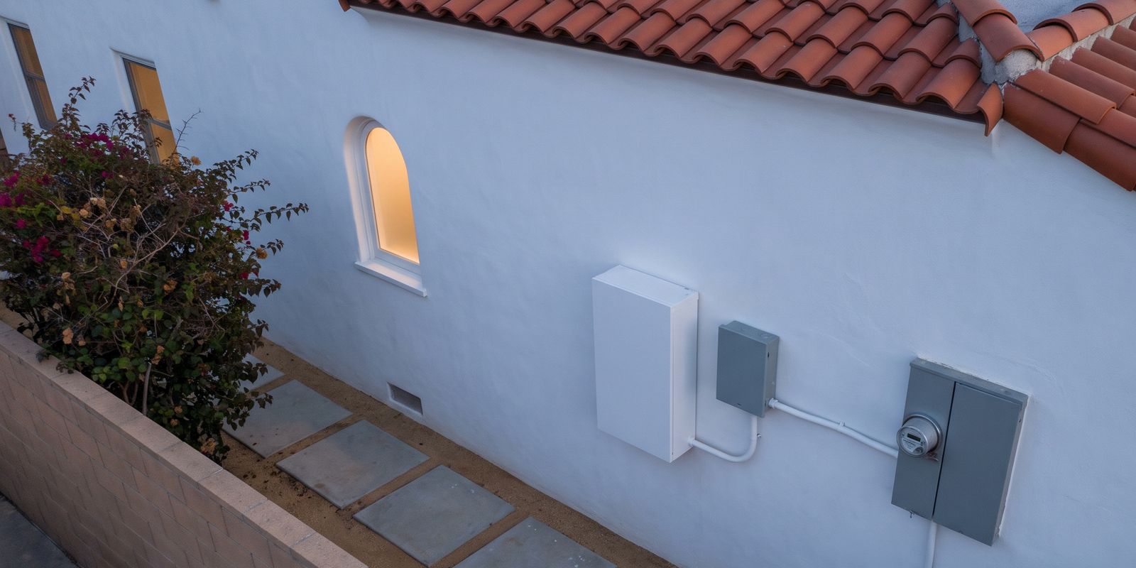 A white home battery and a gray gateway box on the white stucco side wall of a Spanish-style Los Angeles house at dusk, seen from above, with conduit running to the meter under the clay tile eave.